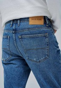 Plave traperice imaju klasični dizajn s dva stražnja džepa, kontrastnim šavovima i kožnom oznakom s natpisom "SALSA JEANS" na pojasu.