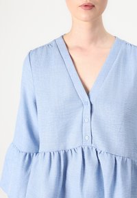 Lyseblå bluse med V-hals, en rad knapper og flagrende ærmer med samlet detalj ved taljen. Bløtt, strukturert stoff.