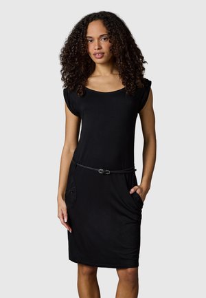 Kobieta z kręconymi włosami, ubrana w czarną sukienkę bez rękawów z cienkim paskiem i rękami w bocznych kieszeniach, stoi na jednolitym tle.