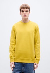 Κίτρινο φούτερ με στρογγυ λή λαιμόκοψη και μικρό λογότυπο. Διαθέτει μακριά μανίκια και ριμπ μανσέτες, σε ουδέτερο φόντο.