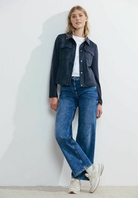Giacca cropped in ecopelle scamosciata blu scuro con due tasche sul petto, abbinata a jeans a gamba larga blu chiaro e sneakers bianche. Texture morbida.