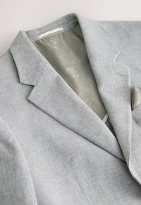 Blazer gris clair avec col à cran, doublure intérieure à motif floral et pochette assortie sur un fond clair.