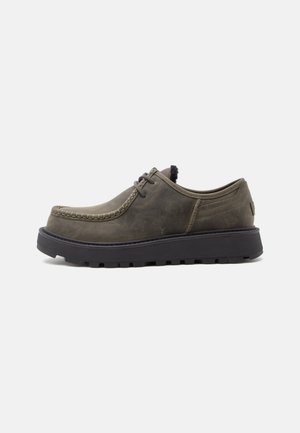 Zapato de piel verde oliva oscuro con cordones, suela negra gruesa y costuras de estilo mocasín a lo largo de la puntera, mostrado de perfil sobre fondo blanco.