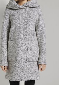 Žena v texturovaném šedém kabátu s kapucí a velkými předními kapsami, stojící na jednorázovém světle šedém pozadí.