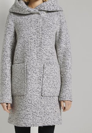 Femme portant un manteau à capuche texturé gris avec de grandes poches avant, se tenant contre un fond gris clair uni.