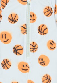 Une combinaison en tissu blanc avec un motif de ballons de basket orange et de visages souriants. Elle dispose d'une fermeture éclair centrale et d'une texture douce.