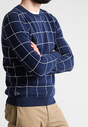 Homme aux bras croisés portant un pull bleu marine à fines lignes blanches en forme de grille et un pantalon beige, debout devant un fond clair uni.