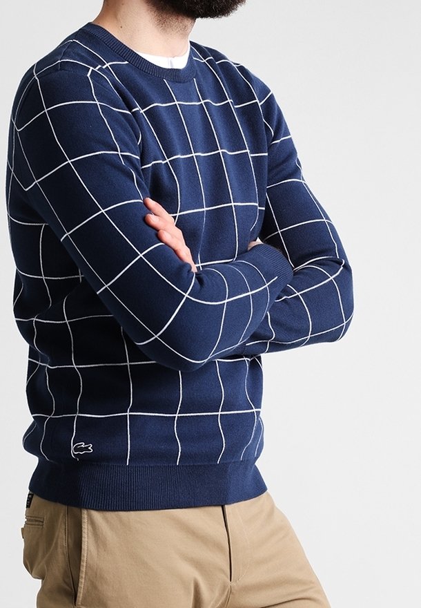 Homme aux bras croisés portant un pull bleu marine à fines lignes blanches en forme de grille et un pantalon beige, debout devant un fond clair uni.