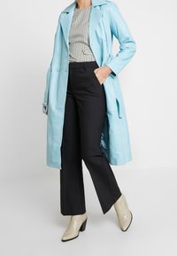 Trench-coat en cuir bleu clair porté sur un haut à carreaux noir et blanc, associé à un pantalon noir et des bottines à texture métallique.