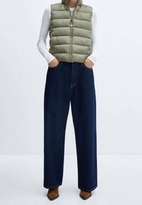 Zielona oliwkowa kamizelka puchowa z zamkiem z przodu, w zestawieniu z luźnymi, ciemnoniebieskimi dżinsowymi spodniami o szerokich nogawkach oraz brązowymi, szpilkowymi botkami.