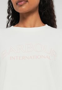 Sweatshirt branco com um grande texto em rosa claro "BARBOUR INTERNATIONAL" atravessando o peito, com decote arredondado e textura suave.