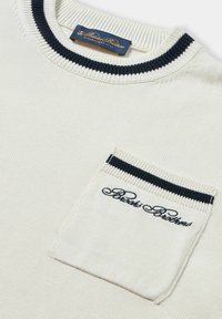 Bílý pletený svetr s krémovým límcem a kapsou lemovanou námořnickou modří. Na kapse je vyšitý logo. Hladký materiál, ležérní design.