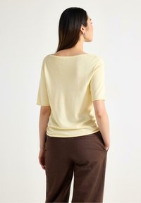 Lys gul kortærmet top med en bred halsudskæring, lavet af et blødt materiale, parret med brune løstsiddende bukser. Simpelt, sømløst design.