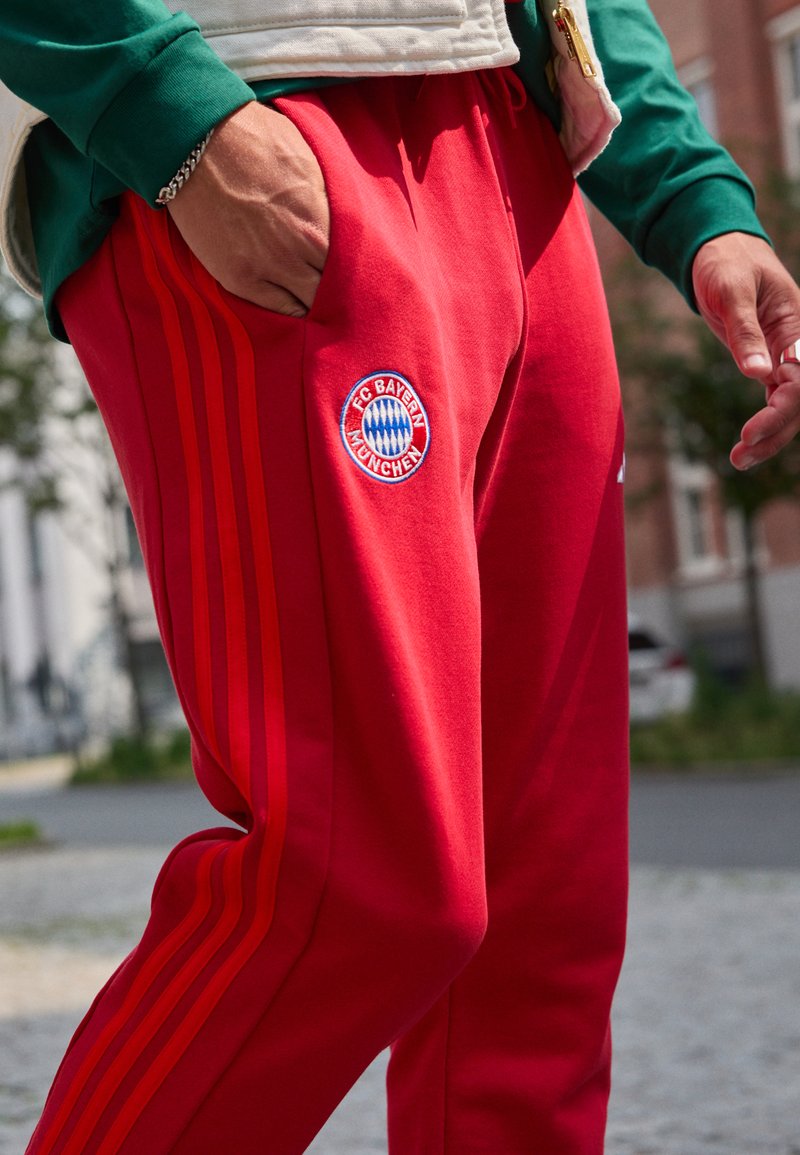 Røde joggebukser har tre røde striper langs sidene, en myk tekstur og en brodert FC Bayern München-logo på låret.