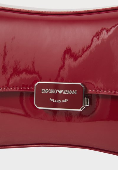 Rød patent lærveske med en blank overflate, som har en sølvmetalllogo plate med teksten "EMPORIO ARMANI MILANO 1981."