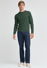Dunkelgrüner Pullover mit Zopfmuster und Rundhalsausschnitt, kombiniert mit dunklen Denim-Jeans und hellen Turnschuhen, präsentiert vor neutralem Hintergrund.