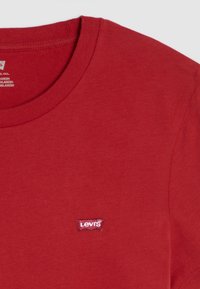 T-shirt en coton rouge avec un col rond, arborant un petit logo Levi's brodé dans le coin supérieur gauche. Texture lisse, design décontracté.
