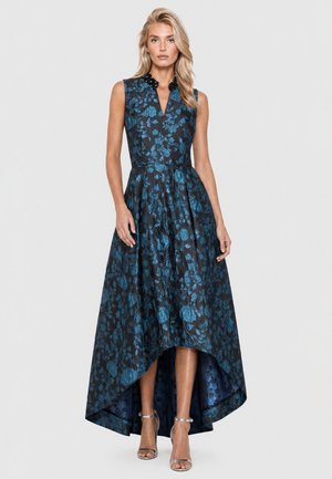 Frau in ärmellosem schwarzem Kleid mit blauem Blumenmuster, hohem-tiefem Saum, schwarzem verziertem Kragen und silbernen Knöchelriemen-Absätzen vor weißem Hintergrund.
