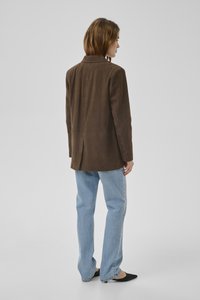 Vrouw met een bruin suède blazer, lichtblauwe jeans en zwarte puntige platte schoenen, zijwaarts staand tegen een effen lichtgrijze achtergrond.