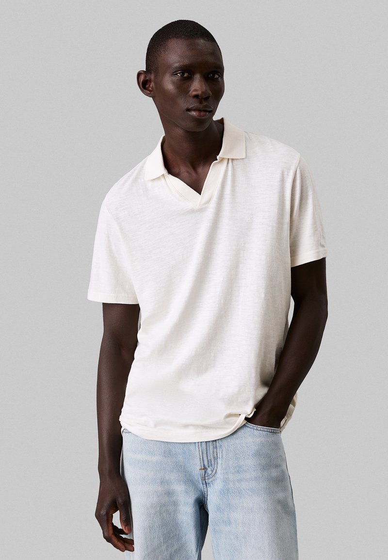 Lichtbeige polo shirt met een gestructureerde afwerking, korte mouwen en een V-hals. Gecombineerd met lichtblauwe denim jeans.