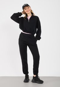 Μαύρο φούτερ με φερμουάρ και ελαστική μέση, συνδυασμένο με μαύρο παντελόνι φόρμας. Φορεμένο με μαύρα αθλητικά παπούτσια και μαύρο καπέλο με λογότυπο.