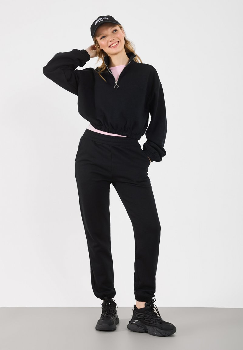 Μαύρο φούτερ με φερμουάρ και ελαστική μέση, συνδυασμένο με μαύρο παντελόνι φόρμας. Φορεμένο με μαύρα αθλητικά παπούτσια και μαύρο καπέλο με λογότυπο.