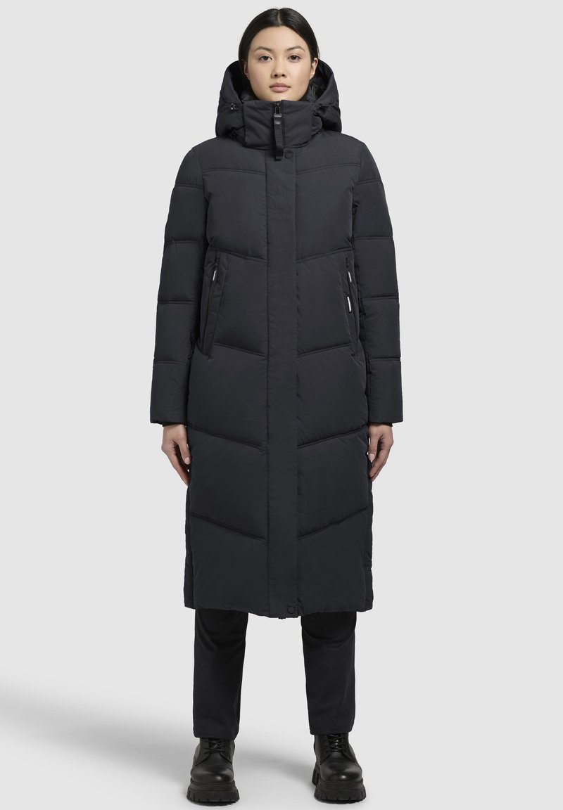 Abrigo de plumas negro largo con capucha, que presenta un cierre con cremallera en la parte delantera, diseño de costillas verticales y dos bolsillos laterales con cremallera. Textura suave.