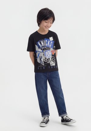 Garçon aux cheveux noirs courts, portant un t-shirt graphique noir des New York Knicks, un jean bleu et des baskets montantes noires, debout les mains derrière le dos.