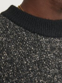 Strikkegenser med en svart ribbestrikket krage, som har et teksturert mønster av svart, grått og kremfargede garn. Nærbilde av stoffet.
