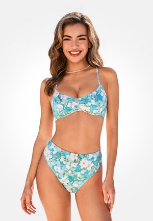 Mujer sonriente con cabello ondulado, usando un bikini azul con estampado floral, bragas de talle alto y tirantes delgados, de pie frente a un fondo blanco liso.