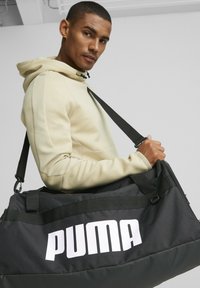 Bolsa de deporte negra con un gran logo blanco de "PUMA", hecha de una tela duradera con un acabado texturizado. Cuenta con asas resistentes y una correa ajustable.