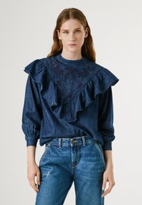 Camicia in denim con un grande colletto arricciato, tessuto di colore blu scuro e un delicato motivo floreale. Maniche lunghe con polsini arricciati, abbinata a jeans blu.