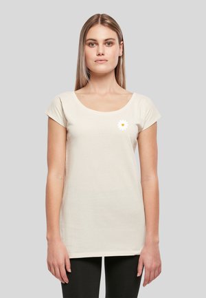 Femme blonde portant un haut beige clair uni à manches courtes avec un petit motif de fleur de marguerite blanche près de la poitrine gauche, debout contre un fond gris.