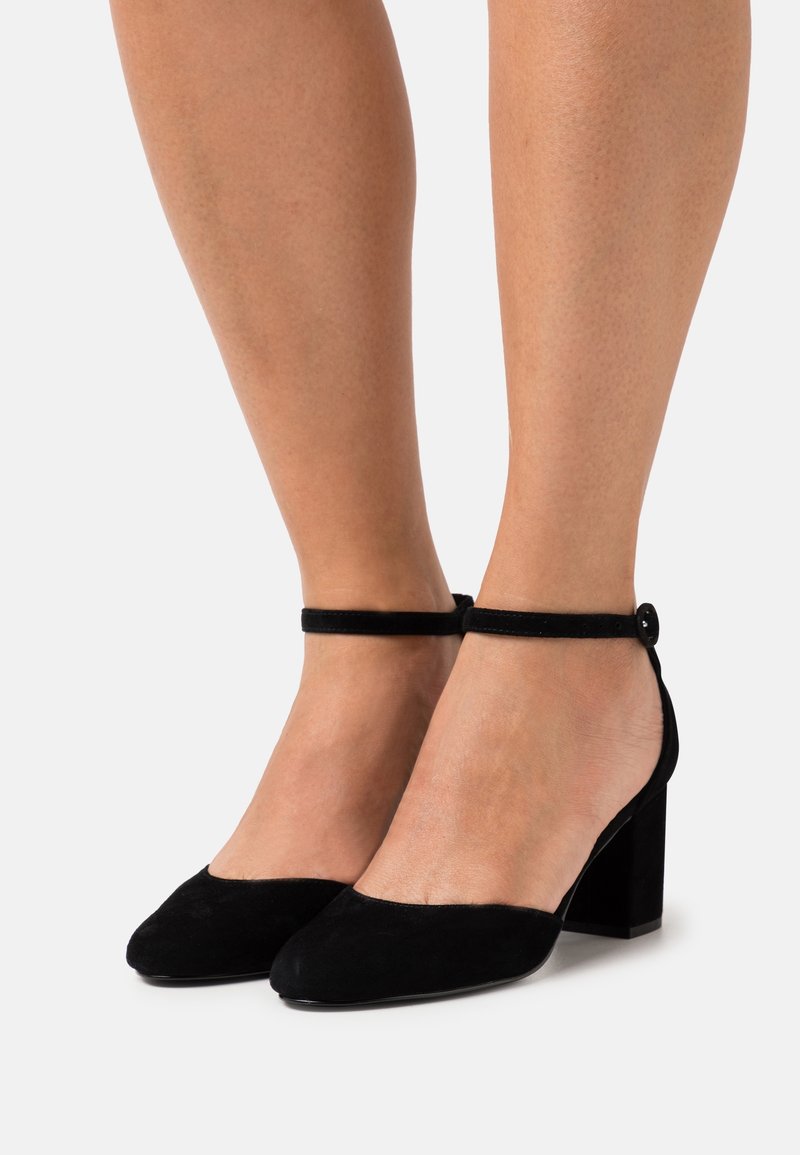 Jambes portant des chaussures en daim noir à talons moyens avec brides à la cheville et boucle ronde, posées sur un fond blanc.