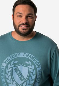 Felpa color turchese con un logo circolare riportante il testo "SPIRIT VICTORY" e "1997", realizzata in materiale morbido con un comodo collo a giro.