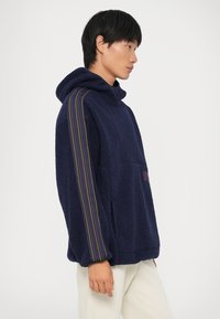Pullover de fleece azul-marinho com capuz, apresentando bolsos laterais, detalhes às riscas em roxo e amarelo nas mangas, superfície texturizada e corte descontraído.