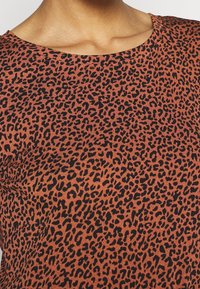 Ruosteenoranssi kangas mustalla leopardikuvioinnilla; näkyvissä pyöreä kaula-aukko. Kangas näyttää sileältä ja istuu tiiviisti.