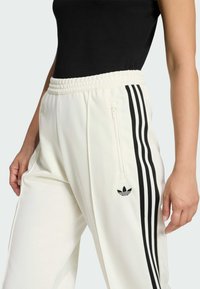 Biele športové nohavice s rebrovaným elastickým pásom, s čiernymi pruhmi na bokoch a malým čiernym logom na dolnej časti nohy.