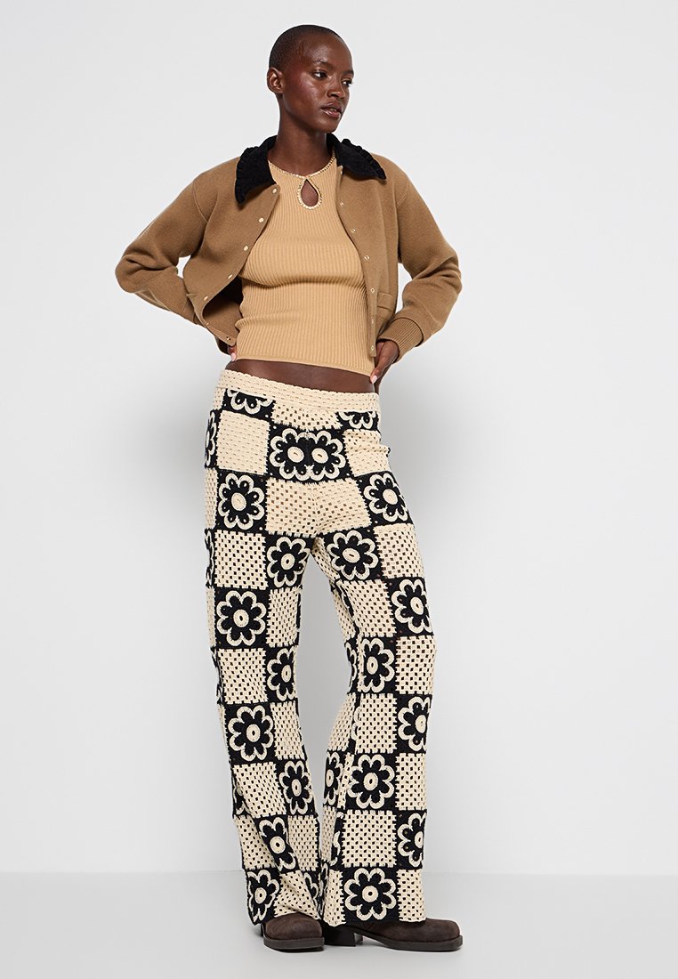 Beiger gerippter Body unter einer braunen Strickjacke mit schwarzem Kragen. Ausgestellte Hosen mit cremefarbenem und schwarzem floralen Häkelmuster. Braune Schuhe.
