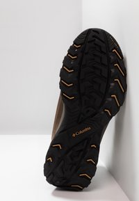 Svart gummisula med strukturerat mönster, med en logotyp i guld. Den övre delen är beige med slät yta.