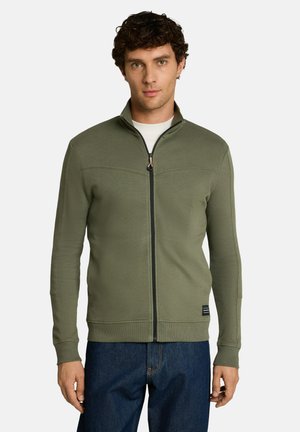 Homme portant une veste zippée vert olive sur une chemise blanche avec un jean bleu foncé, debout sur fond blanc.