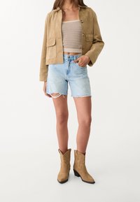 Veste en daim marron, top court beige à côtes, shorts en denim léger délavé et bottes de cowboy beiges avec des détails brodés.