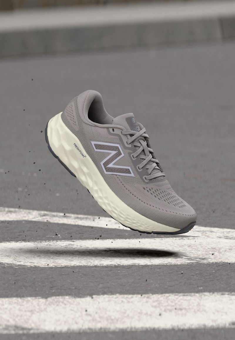 Γκρι αθλητικό παπούτσι New Balance με παχιά λευκή σόλα σε αέρα πάνω από άσφαλτο με λευκές λωρίδες δρόμου.