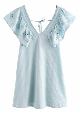 Femme en robe courte bleu clair avec manches à volants et sandales blanches, debout près d'une table et de chaises blanches d'extérieur sur un pavé en pierre.