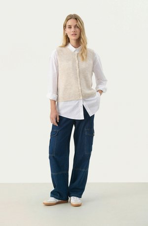 Béžová texturovaná bezrukávová vesta přes bílou košilovou košili, doplněná modrými širokými cargo kalhotami. Bílé tenisky dotvářejí outfit.