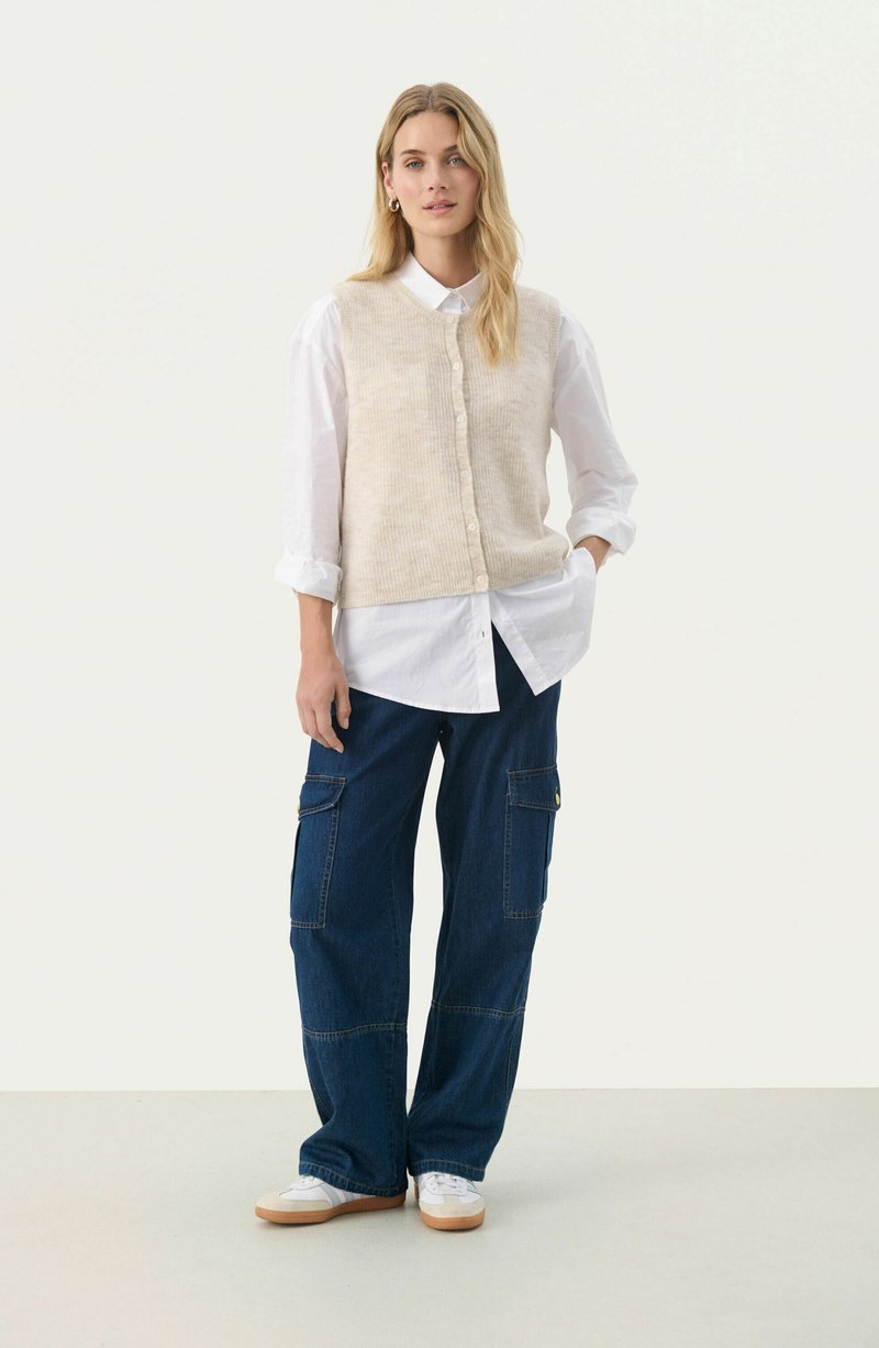 Beige texturerad ärmlös väst över en vit skjorta med knappar, i kombination med blå vida cargo-byxor. Vita sneakers fulländar outfiten.