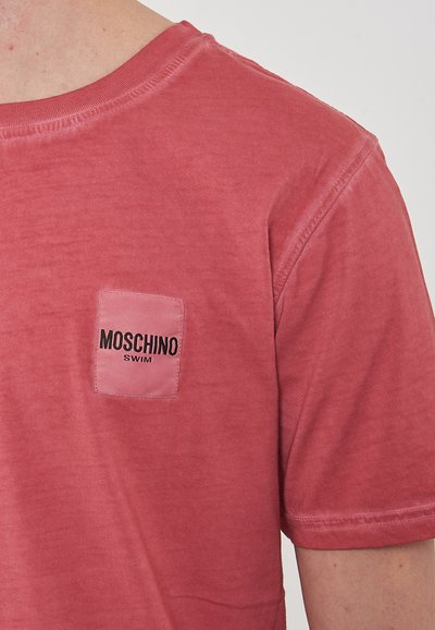 T-shirt in cotone rosso con collo a giro e maniche corte, caratterizzato da un patch quadrato sul petto con la scritta "MOSCHINO SWIM" in lettere nere.