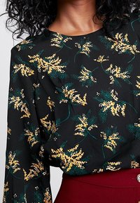 Musta pitkähihaine paita, jossa on keltainen kukkakuosi, käytetään tunkattuna korkeavyötäröisiin burgundinpunaisiin housuihin, joissa on nappiyksityiskohta.