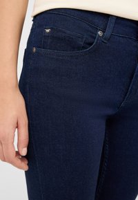 Ciemnoniebieskie jeansy z dżinsu o przylegającym kroju. Posiadają małą przednią kieszeń, srebrne okucia oraz gładką, lekko teksturowaną powierzchnię.