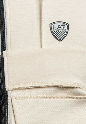 Tessuto sportivo beige con finitura strutturata, caratterizzato da una zip nera e un logo a scudo. Il design include un dettaglio di tasca orizzontale.